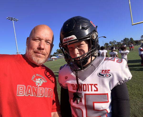 Chris Naylon in a red Port Charlotte Bandits shirt next to his son Sam, wearing his Bandits number 55 jersey and helmet, on the sideline.